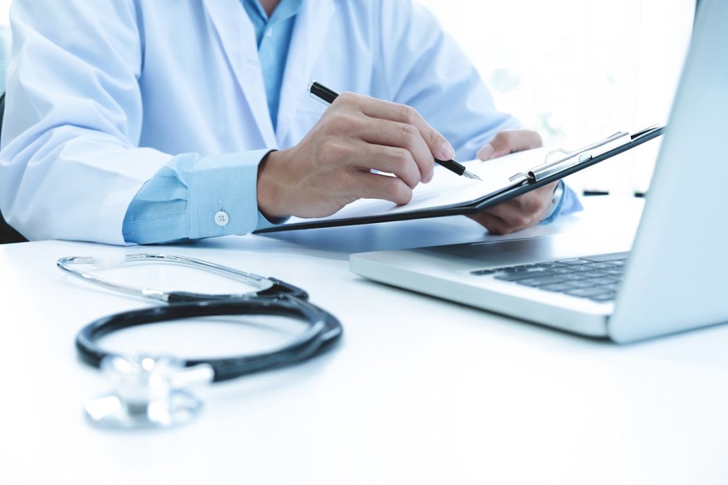 Doctor working with laptop computer and writing on paperwork. Ho Doctor working with laptop computer and writing on paperwork. Hospital background.